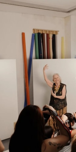 Une femme âgée souriante pose avec élégance devant des rouleaux de papier de fond dans un studio, photographiée par plusieurs personnes au premier plan.