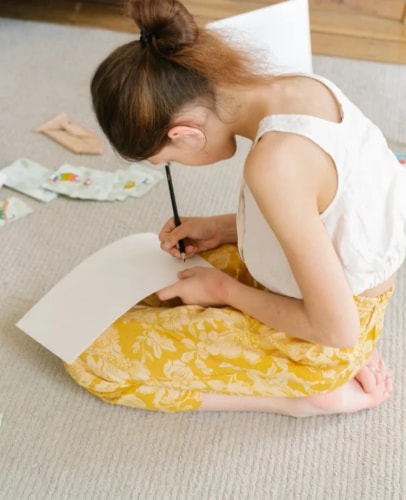 Une jeune fille aux cheveux attachés, assise en tailleur sur un tapis, dessine avec un crayon sur une feuille blanche. Elle porte un haut blanc et un pantalon jaune à motifs floraux.