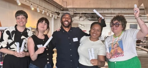 Cinq personnes souriantes posent ensemble en intérieur. Elles tiennent des diplômes roulés ou des petits papiers en main. Elles portent des tenues décontractées et ont l'air heureuses.