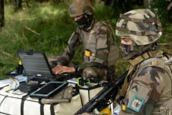 Deux soldats en uniforme camouflé, un assis devant un ordinateur portable et l'autre avec une arme, travaillant ensemble dans un environnement forestier.