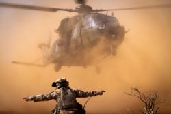 Un hélicoptère militaire survolant une zone poussiéreuse avec un soldat en avant-plan, les bras écartés.