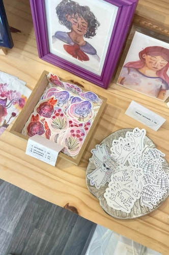 Vue de dessus d'un stand artisanal avec des illustrations encadrées, une boîte remplie d'autocollants colorés à motifs de fleurs et un plateau rond présentant des autocollants en forme de chats blancs.