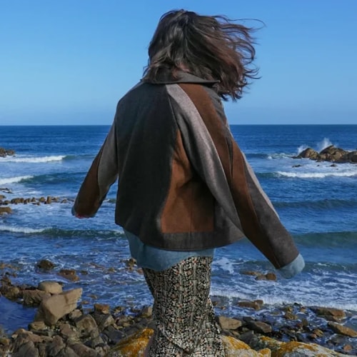 Personne aux cheveux longs marchant vers la mer, vêtue d’une veste sombre et d’un pantalon à motifs, sur une plage rocheuse sous un ciel bleu.