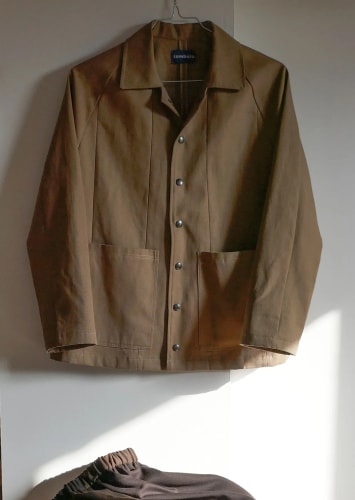 Veste légère marron en cuir ou tissu satiné, à col classique, manches longues et boutons sur le devant, suspendue à un cintre.
