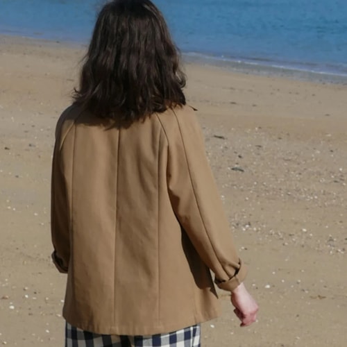 Une personne aux cheveux longs, vue de dos, marche sur une plage en portant une veste beige et un pantalon à carreaux.