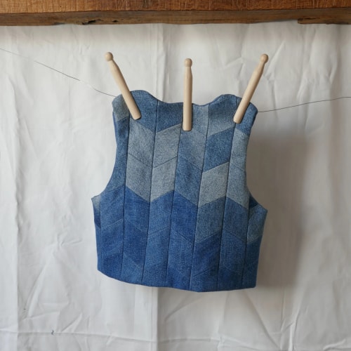 Gilet sans manches tricoté en patchwork bleu, suspendu à trois aiguilles en bois sur un fond blanc.