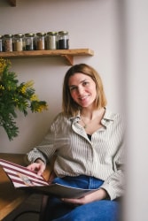 Fanny Taudou souriante assise avec un livre ouvert à la main, portant une chemise à rayures et un jean, avec une plante verte à fleurs jaunes à gauche et une étagère en bois avec des bocaux contenant diverses graines en arrière-plan.