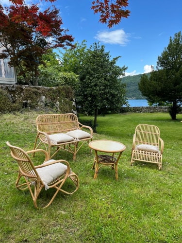 Un ensemble de mobilier de jardin en rotin comprenant un canapé, deux fauteuils et une table, situé sur une pelouse verte, avec des arbres et un plan d'eau en arrière-plan.