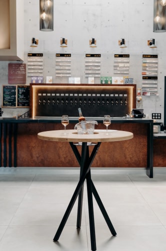 Un bar moderne avec un comptoir sombre, des murs en béton et un éclairage doux. Au centre, une table ronde en bois avec un seau à glace contenant une bouteille et deux verres de vin. À l'arrière-plan, une sélection de boissons est affichée sur un mur.