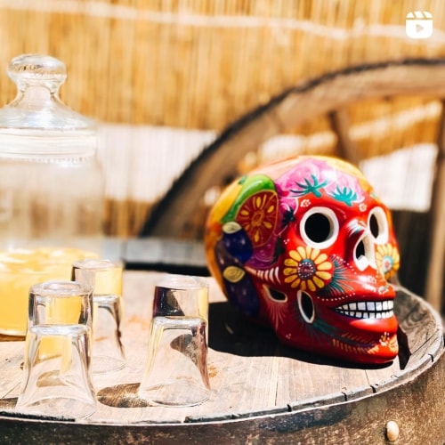 Un crâne coloré de style mexicain avec des motifs floraux, posé sur un fût en bois, à côté de plusieurs petits verres transparents et d'un récipient en verre.