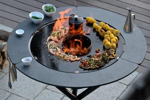 Gril circulaire avec des crevettes marinées, du maïs grillé et des légumes colorés, entouré de bols d'herbes fraîches et d'un pot noir au centre, avec des flammes.