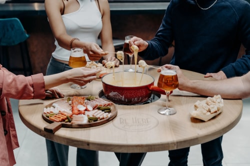 Groupe de personnes partageant une fondue, avec des verres de bière et une planche de charcuterie sur une table en bois.