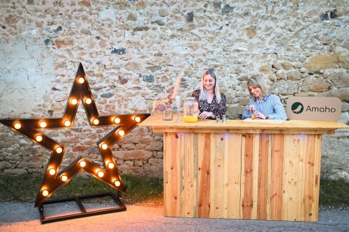 Deux femmes souriantes se tiennent derrière un comptoir en bois, préparant des boissons; à gauche se trouve une grande étoile lumineuse, et à droite, un panneau avec le logo "Amaho". En arrière-plan, un mur en pierre.