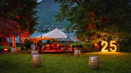 Une terrasse élégante éclairée par des guirlandes lumineuses, où des invités se rassemblent, entourée d'arbres, de fût en bois et d'un grand panneau lumineux avec le nombre 25, le tout avec un lac et des montagnes en arrière-plan.