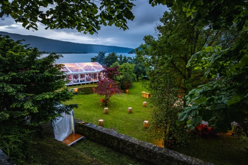 Vue panoramique d'un jardin avec une tente transparente éclairée, entourée de verdure et d'un lac en arrière-plan sous un ciel nuageux.