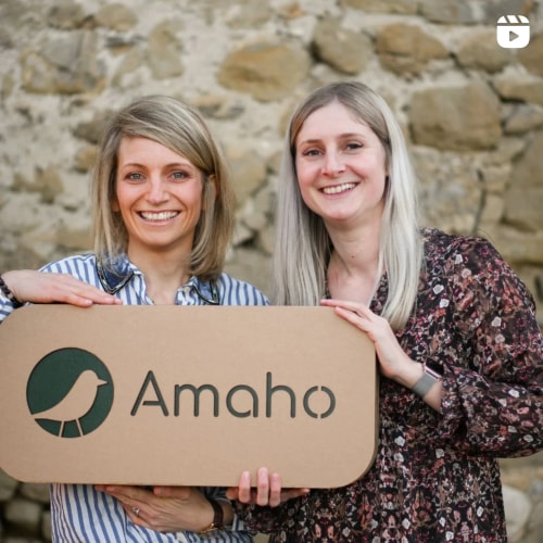 Deux femmes souriantes tenant une grande pancarte avec le logo et le nom 'Amaho' dans un environnement naturel.