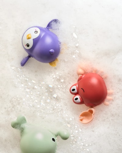 Trois jouets en plastique dans une baignoire pleine de mousse : un pingouin violet, un crabe rouge et une grenouille verte.