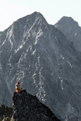 Une personne avec une veste orange est assise sur un rocher escarpé et contemple une immense montagne rocheuse sous un ciel clair.