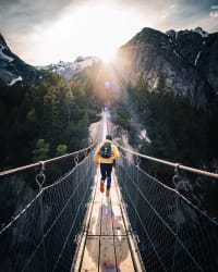 Une personne portant une veste jaune et un sac à dos marche sur un pont suspendu vers des montagnes sous un soleil éclatant.
