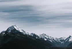 Chaîne de montagnes majestueuse aux sommets enneigés se découpant sur un ciel nuageux et dramatique dans des tons gris et bleus.