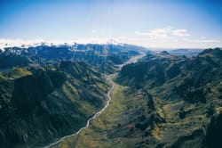 Vue aérienne d'une vallée verdoyante islandaise traversée par une rivière, entourée de montagnes rocheuses et de sommets enneigés sous un ciel bleu dégagé.