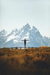 Une silhouette debout sur une barrière, les bras étendus, face à une haute montagne enneigée dans un paysage de prairie automnale.