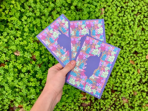 Main tenant trois cartes ou enveloppes colorées à motifs floraux au-dessus d’un tapis de petites feuilles vertes.