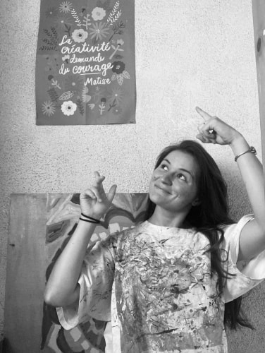 Jeune femme souriante pointant vers une affiche fleurie avec une citation en français, dans une pièce aux murs clairs.