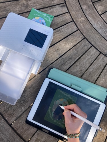 Deux appareils électroniques ouverts sont posés sur une terrasse en bois : une liseuse blanche et une tablette affichant une carte, sur laquelle une main écrit au stylet.