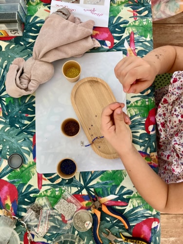 Une personne peint un petit objet en bois avec un pinceau, entourée de pots de peinture et de tissus colorés à motifs tropicaux.