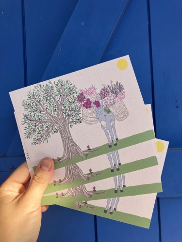 Main tenant des cartes illustrées montrant un arbre coloré et plusieurs personnages, posées sur un fond bleu.