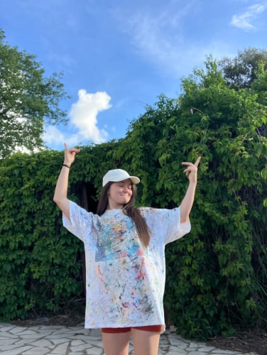Une femme souriante, vêtue d’une robe ample colorée, lève les bras devant une haie verdoyante, sous un ciel bleu parsemé de nuages.