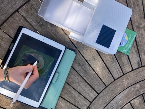 Une personne dessine sur une tablette affichant une carte, posée sur un étui bleu, à côté d’une boîte de rangement ouverte sur une terrasse en bois.