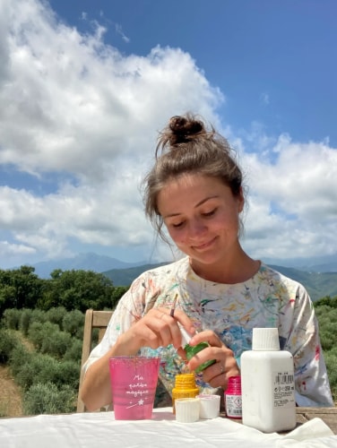 Une jeune femme prépare des produits cosmétiques sur une terrasse, entourée d’oliviers et de montagnes sous un ciel nuageux.