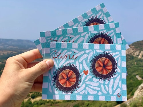 Main tenant trois cartes illustrées turquoise, ornées de fleurs rouges et de motifs blancs, avec le mot « Lumi », devant un paysage montagneux.