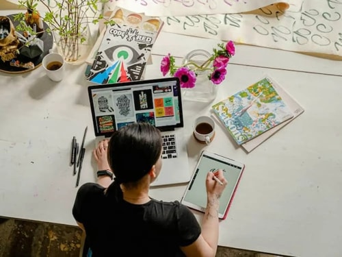 Une vue aérienne d'une personne travaillant sur un ordinateur portable sur une table avec des stylos, une tasse de café et un carnet de croquis. Des fleurs et des magazines décorent la table.