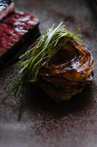 Un plat gourmet mettant en vedette un oignon enroulé grillé, garni de brins d'herbes vertes, à côté d'un morceau de viande rouge servie sur une assiette sombre.