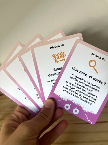 Main tenant une éventail de cartes pédagogiques en français, dont une carte expliquant une note après un achat et une autre sur le biais de confirmation.