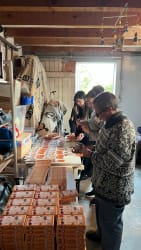 Trois personnes trient et organisent des piles de petits paquets orange sur une longue table dans une pièce intérieure, avec des étagères et des tissus en arrière-plan.