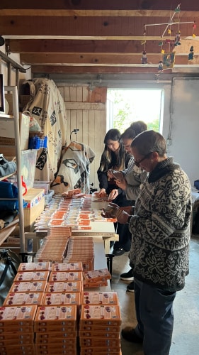 Trois personnes trient et organisent des piles de petits paquets orange sur une longue table dans une pièce intérieure, avec des étagères et des tissus en arrière-plan.