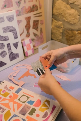 Une personne crée un collage artistique sur une table, manipulant des formes en papier découpé colorées, avec des œuvres abstraites en arrière-plan.