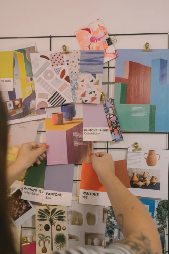 Une main tient plusieurs feuilles de papier imprimées avec des motifs colorés abstraits, floraux et géométriques sur une table blanche.
