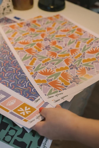 Une main tient des feuilles de papier imprimées avec divers motifs colorés, géométriques et floraux sur un bureau.