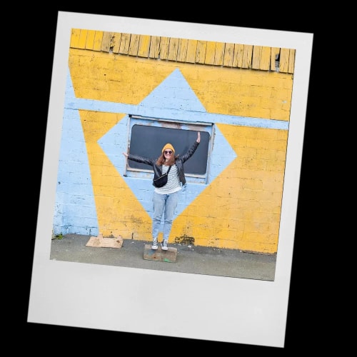 Portrait d'une femme souriante aux cheveux bruns devant un mur décoré de formes géométriques colorées.