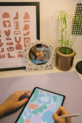 Une personne dessine sur une tablette numérique avec un stylet sur un bureau, reflétée dans un miroir décoratif en terrazzo. Une plante en pot et un cadre abstrait complètent la scène.