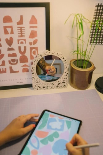 Une personne dessine sur une tablette numérique avec un stylet sur un bureau, reflétée dans un miroir décoratif en terrazzo. Une plante en pot et un cadre abstrait complètent la scène.