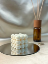 Bougie blanche Fiamme en forme de cube composée de petites sphères, posée sur un miroir rond, avec en arrière-plan un diffuseur d'ambiance en verre brun avec des bâtonnets en bois.