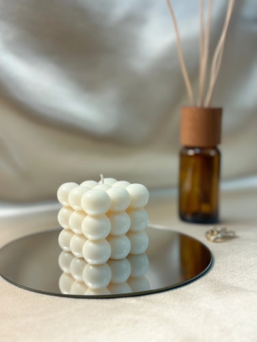 Bougie blanche Fiamme en forme de cube composée de petites sphères, posée sur un miroir rond, avec en arrière-plan un diffuseur d'ambiance en verre brun avec des bâtonnets en bois.