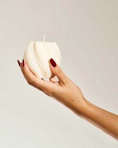 Bougie blanche Fiamme en forme de cube composée de petites sphères, posée sur un miroir circulaire, avec un diffuseur de parfum en verre brun flou en arrière-plan.