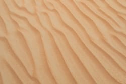 Gros plan sur des vagues de sable formées par le vent sur une plage, montrant une texture ondulée et des nuances de beige.